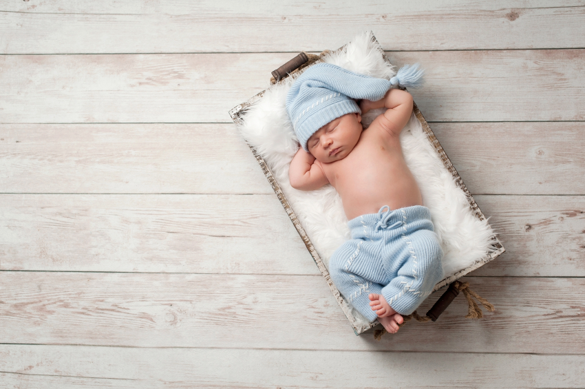 Newborn baby sleeping in a wooden crate on a whitewashed wooden floor. He is wearing light blue, upcycled pajamas with a matching sleeping cap.